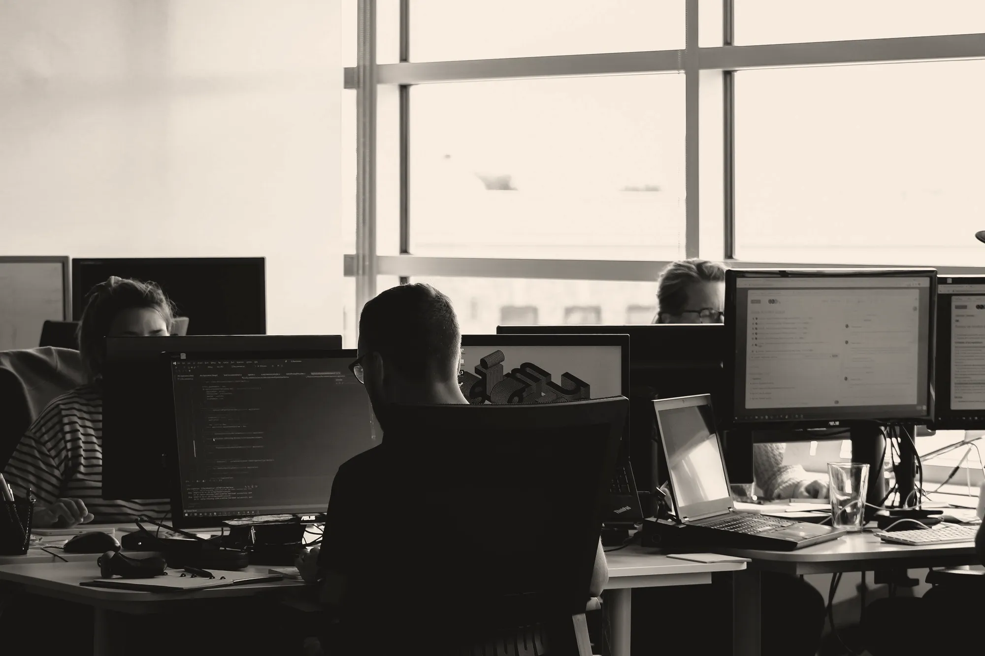 Developers seen from behind, with code on their monitors in a bright office.